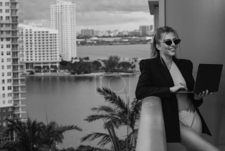 Téléchargez les photos : Femme d'affaires confiante sexy en costume d'affaires de mode en utilisant un ordinateur portable dans le bureau moderne. Miami balcon vue - en image libre de droit
