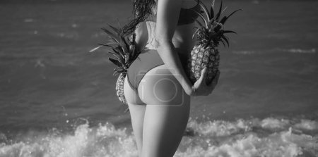 Téléchargez les photos : Womans buttock in bikini. Close up female swimsuit model on adriaticor bali sea bech. Girl holding pineapple near her butt ass. View from back - en image libre de droit