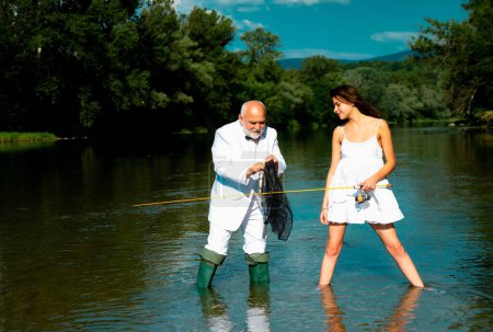 Téléchargez les photos : Couple drôle de jeune jolie femme beauté et vieux pêcheur debout dans la rivière avec canne à pêche. Vieux senior et fille pêche - en image libre de droit