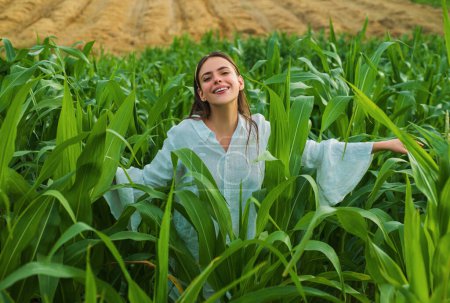 Téléchargez les photos : Jeune fille heureuse montrant le maïs récolté dans le champ. Femme américaine en robe blanche récolte du maïs. Jeune agricultrice à la récolte de maïs - en image libre de droit
