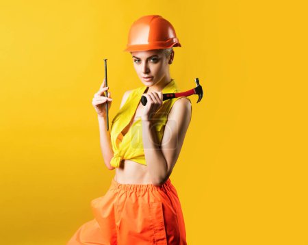 Foto de Mujer sexy en casco para usar martillo. Mujer joven clavando clavos en el taller sobre fondo amarillo aislado. Modelo femenino trabajando en la construcción. Todo puede ser concepto fijo - Imagen libre de derechos