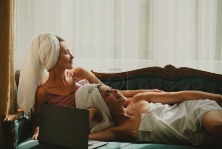 Téléchargez les photos : Jeunes amies détendues avec tasse de café et ordinateur portable au lit à la maison. Joyeux fille sexy avec peignoirs serviettes sur la tête dans la chambre après le réveil du matin - en image libre de droit