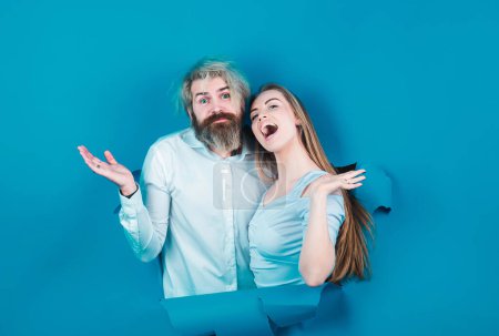 Photo for Surprised couple of bearded man and happy woman making hole in paper looking through hole. Advertising board, blue background. Couple through hole in blue paper, copy space for text or slogan - Royalty Free Image