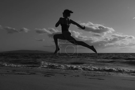 Téléchargez les photos : Une femme court sur la plage. Fille sportive active courant le long de l'océan surf près de la piscine d'eau pour garder la forme et la santé. Fitness femme, entraînement de jogging et activité sportive pendant les vacances d'été - en image libre de droit