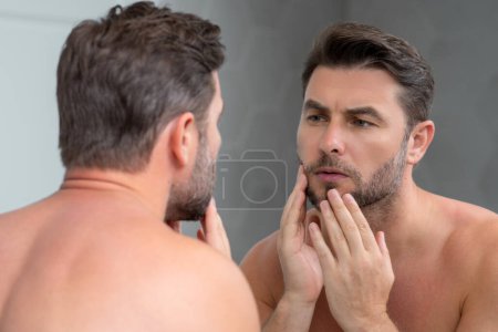 Millennial hispanischer Mann, der in Spiegel, Gesichtshaut und Stoppeln blickt. Männliche Schönheitspflege. Hautpflege, home spa. Schönheitsporträt eines schönen Mannes. Spa-Modell, feuchtigkeitsspendende Pflegecreme