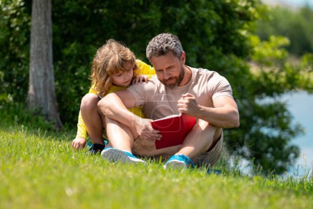 Téléchargez les photos : Père et fils assis sur l'herbe verte dans le jardin et le livre de lecture ensemble. Joyeux livre de lecture en famille ensemble dans la nature verte. Père est assis avec son fils lire le livre. Le gamin est très intéressé. - en image libre de droit