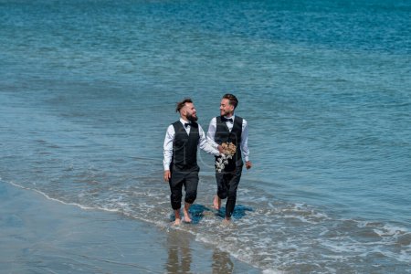 Foto de Los novios gay caminando juntos en la playa del mar durante el día de la boda. Hombres románticos en el agua de mar. Boda de pareja gay. Pareja homosexual gay, pareja LBGT en ceremonia de boda, LGBTQ - Imagen libre de derechos