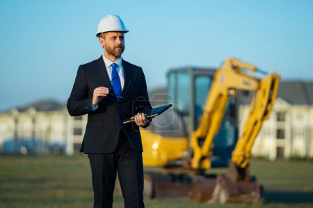 Photo for Civil engineer worker at a construction site. Mature engineer worker. Man in suit and hardhat helmet at construction site. Middle aged head civil engineer worker standing outside near excavator - Royalty Free Image