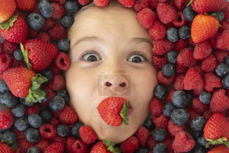 Téléchargez les photos : Visage d'enfant avec baies en gros plan. Mélange de fraises, bleuets, framboises, mûres. Mélange assorti de fraises, bleuets, framboises, mûres. Les enfants drôles font face de près - en image libre de droit