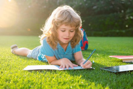 Téléchargez les photos : Garçon mignon avec écriture au crayon sur un cahier posé sur de l'herbe. Enfant lisant un livre dans le parc d'été - en image libre de droit