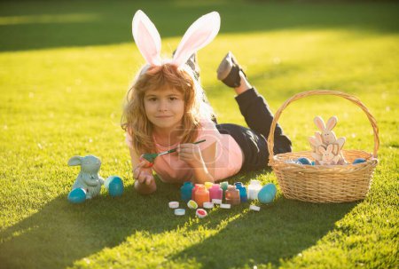 Foto de Niño con traje de conejo con orejas de conejo pintando huevos de Pascua en la hierba en el parque de primavera - Imagen libre de derechos