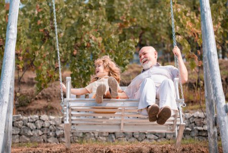 Photo for Old grandfather and young child grandson swinging in garden outdoors. Grand dad and grandson sitting on swing in park - Royalty Free Image
