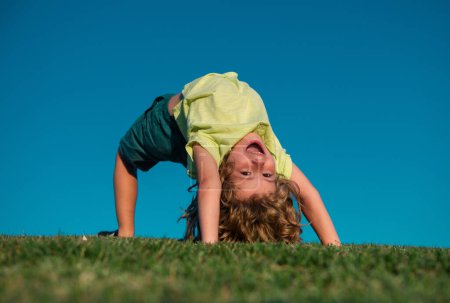 Téléchargez les photos : Portrait d'un enfant souriant à l'envers sur herbe verte. Petit garçon mignon profitant de la nature en plein air. Enfant en bonne santé jouant dehors dans le parc d'été. Sport actif en plein air sain. Activité amusante. - en image libre de droit