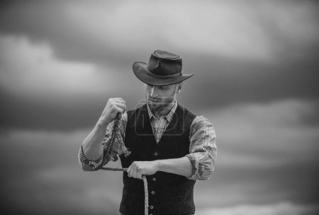 Foto de Un vaquero. Propietario del rancho. Campesino y concepto agrícola. Hombre guapo con sombrero de vaquero y atuendo de estilo rústico. Vaquero con cuerda de lazo en el fondo del cielo - Imagen libre de derechos