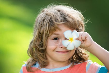 Téléchargez les photos : Portrait d'un enfant heureux riant avec des yeux plumeria fleur. Gros plan positif visage des enfants. Couvre les yeux avec le concept de fleur - en image libre de droit