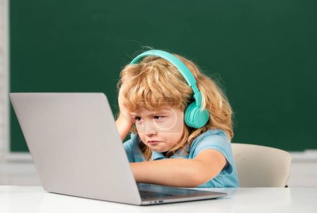 Photo for Angry sad child boy in headphones using a laptop and study online with video call teacher at school. Little funny system administrator or programmer - Royalty Free Image