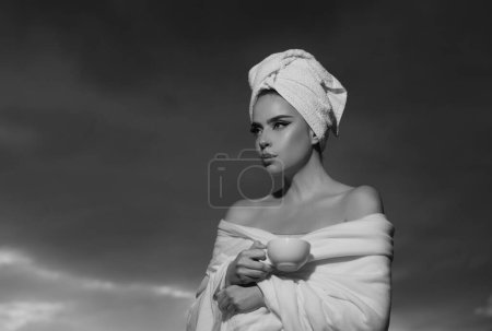Téléchargez les photos : Femme avec serviette sur la tête sentant le café dans la nuit au ciel dramatique en plein air. Spa du matin. Fille avec tasse de café - en image libre de droit