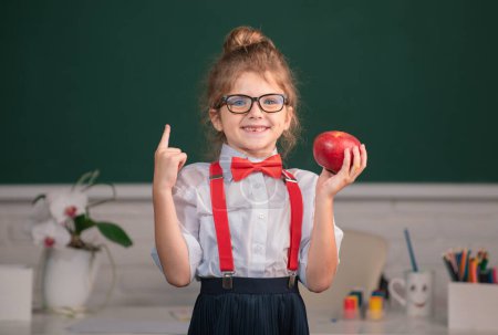 Foto de De vuelta a la escuela. Retrato de colegiala nerd estudiante mantenga manzana con sorprendente expresión apuntando con el dedo contra pizarra - Imagen libre de derechos
