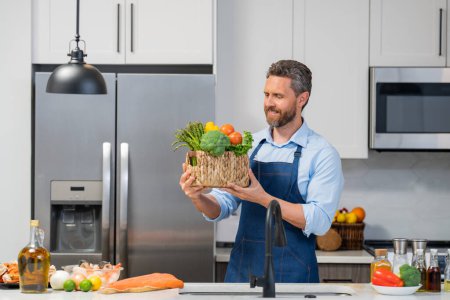 Photo for Chef in cook apron cooking in kitchen. Man on kitchen with vegetables. Portrait of casual man cooking in the kitchen with vegetable ingredients. Guy preparing salad at home in kitchen - Royalty Free Image