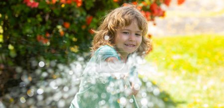 Foto de Niño jugando en el jardín, vierte de la manguera, hace una lluvia. Concepto de infancia feliz. Un niño derramando agua sobre los árboles. Riego infantil de primavera, pancarta - Imagen libre de derechos