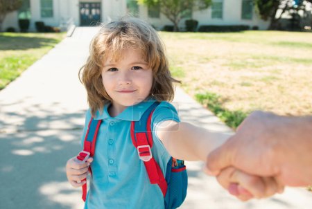 Foto de Niño pequeño con mochila sosteniendo la mano de los padres en el fondo borroso edificio de la escuela. Niño con mochilas parado en el parque cerca de la escuela - Imagen libre de derechos