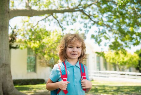 Téléchargez les photos : Écolier avec sac à dos marche sur la pelouse verte. École primaire et éducation. Des enfants de l'école primaire. Portrait d'élève près de l'école - en image libre de droit