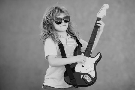 Foto de Retrato de un niño divertido con gafas practicando una canción durante una lección de guitarra en la calle. Concepto musical, escuela de música para niños. Concierto de rock - Imagen libre de derechos