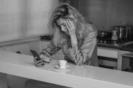 Téléchargez les photos : Triste femme boire du café et en utilisant le téléphone à la maison le matin. Fatigué femme madel bavarder smartphone en pyjama sexy à la cuisine - en image libre de droit