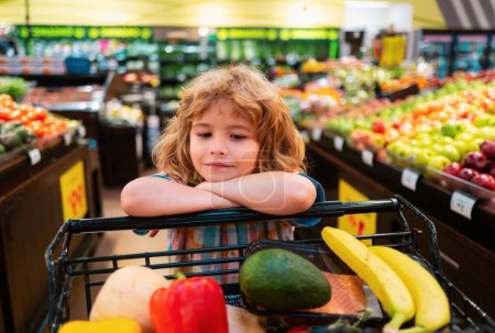 Téléchargez les photos : Enfant garçon fait du shopping dans un supermarché. Petit garçon mignon avec un panier plein de légumes et de fruits biologiques frais dans une épicerie ou un supermarché - en image libre de droit