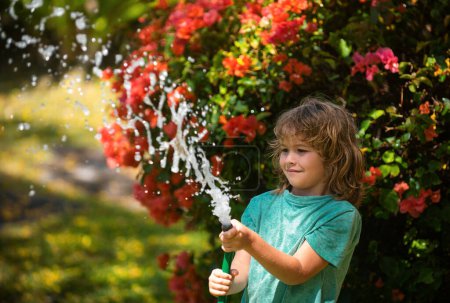 Téléchargez les photos : Un garçon heureux verse de l'eau dans un tuyau. Enfance américaine. Enfant arrosant des fleurs dans le jardin. Jardinage à domicile - en image libre de droit