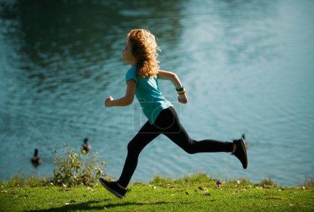 Téléchargez les photos : Les enfants coureurs courent dans le parc. Garçon courant dans le parc en été dans la nature. Sports de plein air et fitness. Enfants courant sur la prairie verte contre la mer ou le lac - en image libre de droit