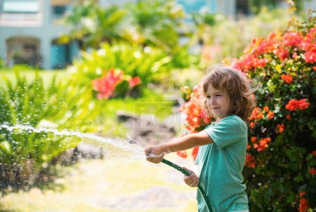 Téléchargez les photos : Mignon petit garçon arrosant des fleurs dans le jardin le jour de l'été. Enfant utilisant un tuyau d'arrosage. drôle enfant arrosage des plantes dans le jardin de la cour - en image libre de droit