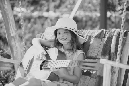Téléchargez les photos : Petit enfant jouant de la guitare. Petite fille à la mode. École de guitare musicale pour enfants. Activité estivale pour les enfants par temps chaud. Petite fille mignonne s'amusant sur une balançoire dans un beau jardin d'été - en image libre de droit
