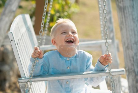 Little smiling boy swinging on a summer sunny day. Child dreaming on swing. Child swinging on playground on sunny summer day in a park. Spring kids portrait. Kid emotions. Portrait of laughing kids