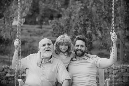 Photo for Grandfather with son and grandson swinging together in summer garden on swing outdoors. Active leisure for man family. Dad, son and granddad hugging - Royalty Free Image