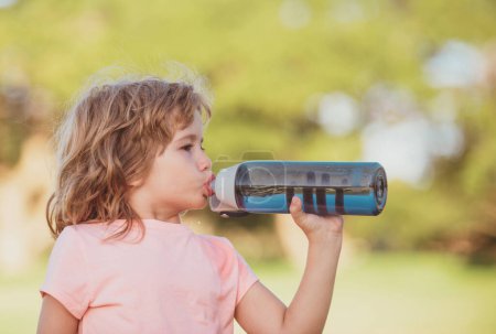 Foto de Lindo niño bebe agua de la botella de deporte en el parque verde. Retrato de primer plano de niño deportivo haciendo ejercicio al aire libre - Imagen libre de derechos