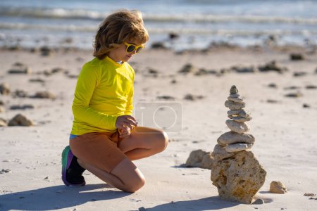 Téléchargez les photos : Enfant avec pyramide de galets en pierre équilibrée sur la plage. Jeu d'enfants avec des pierres zen sur la plage de la mer, méditation, spa, harmonie, calme et équilibre concept pour les enfants - en image libre de droit