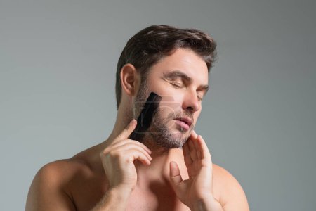 Téléchargez les photos : Homme caucasien essayant de faire un peigne de cheveux de barbe par lui-même avec peigne. Une coupe masculine. Une coupe masculine. Carber shop. Peigne de coiffeur. Concept de coiffure de barbe peignante. Coiffure et peigne pour hommes - en image libre de droit