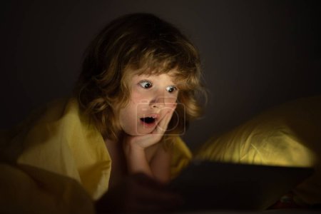 Foto de Niño jugando en una tableta digital. Niño usando tableta para juegos y aprendizaje en línea mientras está acostado en la cama, reflejo de luz de la pantalla en su cara. Niño jugar tableta bajo manta en la noche en una habitación oscura - Imagen libre de derechos