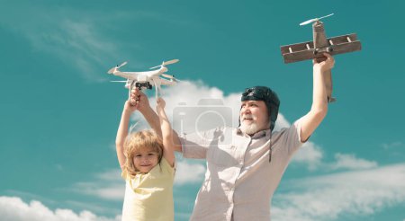 Téléchargez les photos : Vieux grand-père et jeune petit-fils enfant jouant avec avion jouet et quadcopter drone contre ciel. Enfant pilote aviateur avec des rêves d'avion de voyager - en image libre de droit