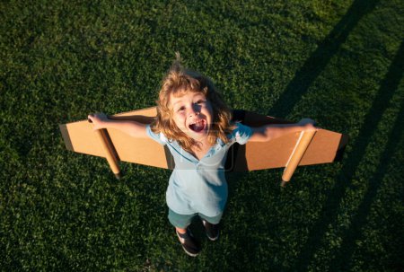 Téléchargez les photos : Incroyable enfant jouant avec des ailes d'avion jouet dans le parc d'été. Technologie d'innovation et concept de réussite. Pilote excité s'amusant sur l'herbe dans le parc. Garçon rêve de voler - en image libre de droit