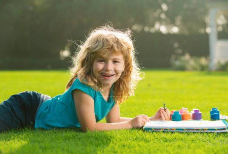 Téléchargez les photos : Jeune artiste peintre talentueux. Enfant garçon dessine dans le parc étendu dans l'herbe en s'amusant sur fond de nature. Enfants croissance créative - en image libre de droit