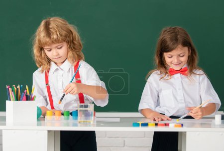 Foto de Los niños de la escuela dibujan dibujos coloridos con lápices de colores en el aula sobre fondo de pizarra. Retrato de alumnos lindos disfrutando de clases de arte y artesanía - Imagen libre de derechos