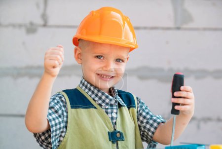 Photo for Happy smiling kid boy twists bolt with screwdriver. Child repairman with repair tool. Child in helmet and boilersuit on construction site. Little worker engineer. Kids builder and repair - Royalty Free Image