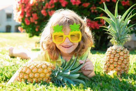 Téléchargez les photos : Petit enfant dans des verres drôles avec des ananas ponte sur l'herbe sur fond de nature d'été - en image libre de droit