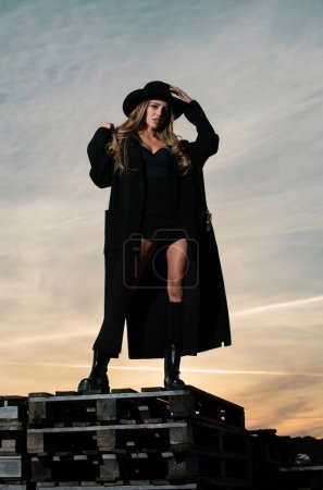 Téléchargez les photos : Femme sexy en manteau de mode noir, chapeau et chaussures noires bottes. Belle jeune femme portant des vêtements de printemps à la mode. Portrait du mannequin en robe tendance - en image libre de droit