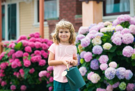 Foto de Lindo niño regando flores con regadera en el jardín. Niño vestido en verano ligero cierra y colorido camiseta, sonriendo y divirtiéndose. Actividades con niños al aire libre - Imagen libre de derechos