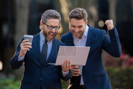 Photo for Two excited business man using laptop outdoor. Colleagues in suit talking outdoor. Two business people talk project strategy. Two american businessmen in suits discuss - Royalty Free Image