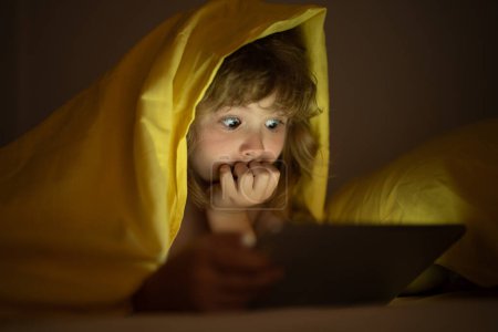 Photo for Kid boy lying on bed and surfing Internet on tablet in dark room. Child using tablet pc at night. Little kid in bed under a blanket looking at the smartphone or tablet at night. Gadget addicted child - Royalty Free Image