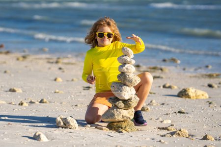 Téléchargez les photos : Méditation estivale pour enfants. Petit enfant jouer avec l'équilibre des pierres pyramidales sur le sable de la plage. Enfant avec pile de pierres sur la mer. Jeu d'enfants avec des pierres équilibre sur la plage. Équilibre et harmonie de vie - en image libre de droit